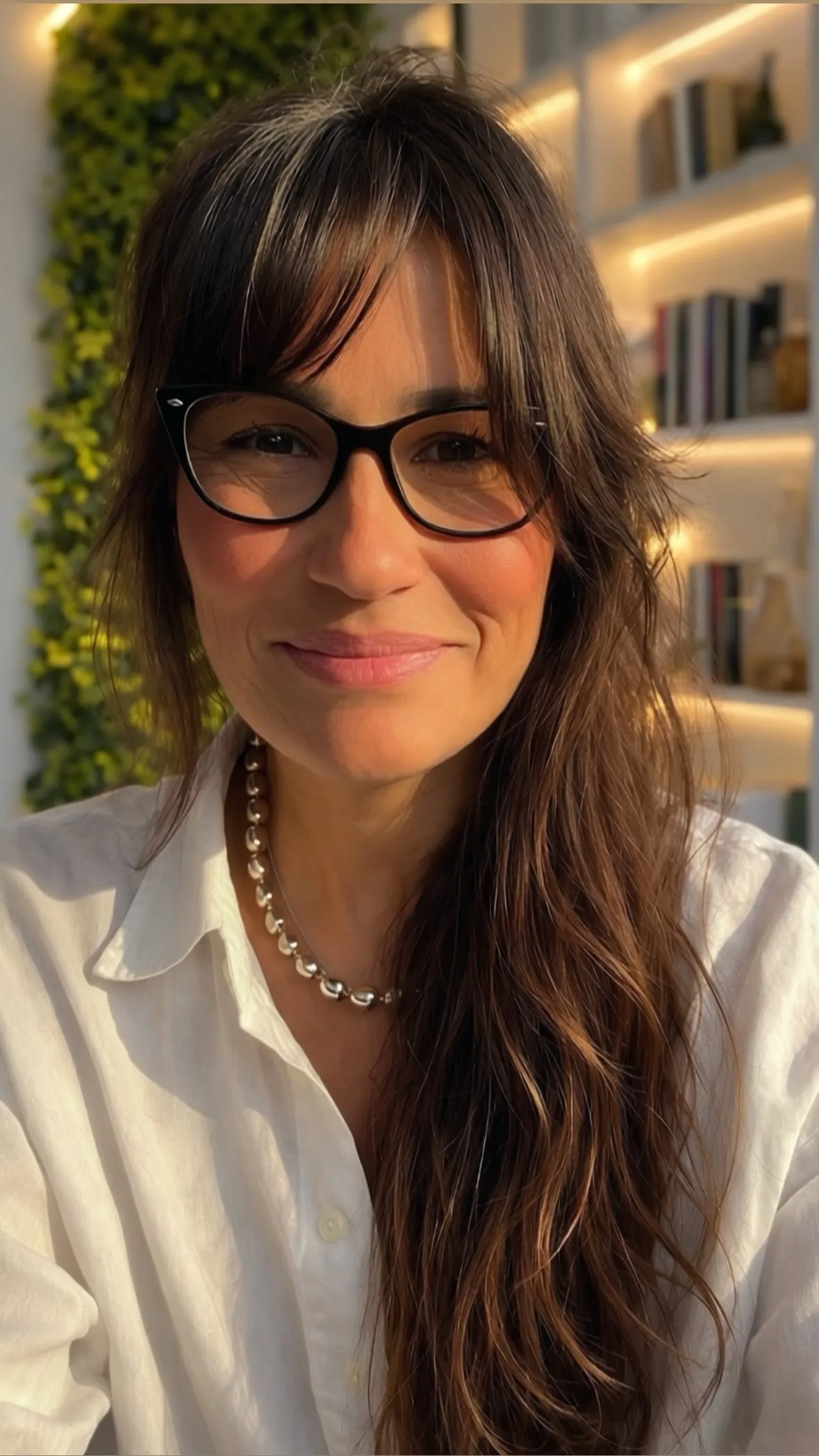 Carolina Mateo sorrindo, de óculos e camisa branca, em frente a uma estante iluminada